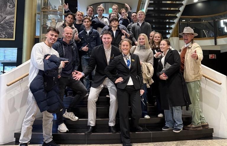 Gruppebilde av en elevklasse og voksne som står samlet i en bred trapp i en hotellobby, smilende og med flere som peker mot kamera